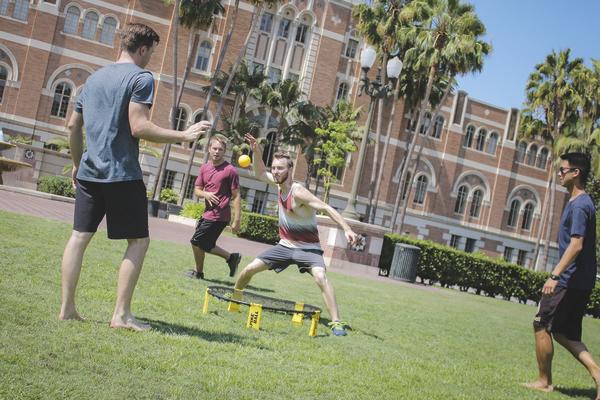 Junge Männer spielen Spikeball auf einer Wiese vor einem historischen Gebäude bei Sonnenschein.