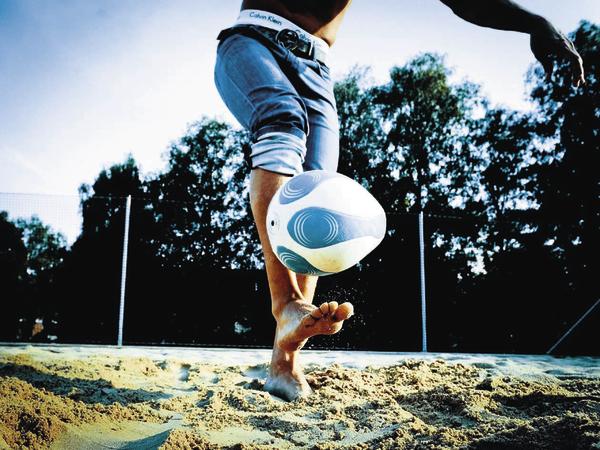 Mann spielt Beachsoccer barfuß im Sand, Ball in der Luft, sonniger Tag im Freien.