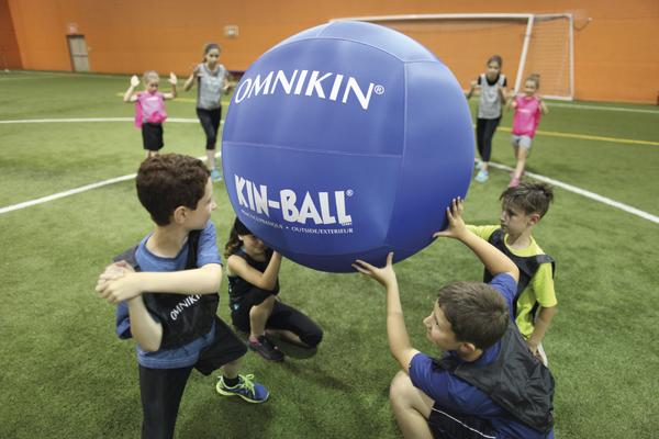 Kinder spielen Kin-Ball mit großem blauen Ball in einer Halle mit grünem Kunstrasen.