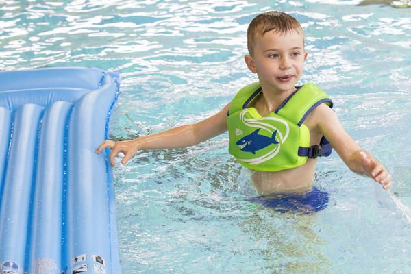 Junge mit grünem Schwimmweste spielt im Pool neben blauem Luftmatratze.