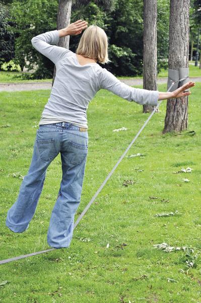 Frau balanciert auf Slackline im Park zwischen Bäumen bei sonnigem Wetter.