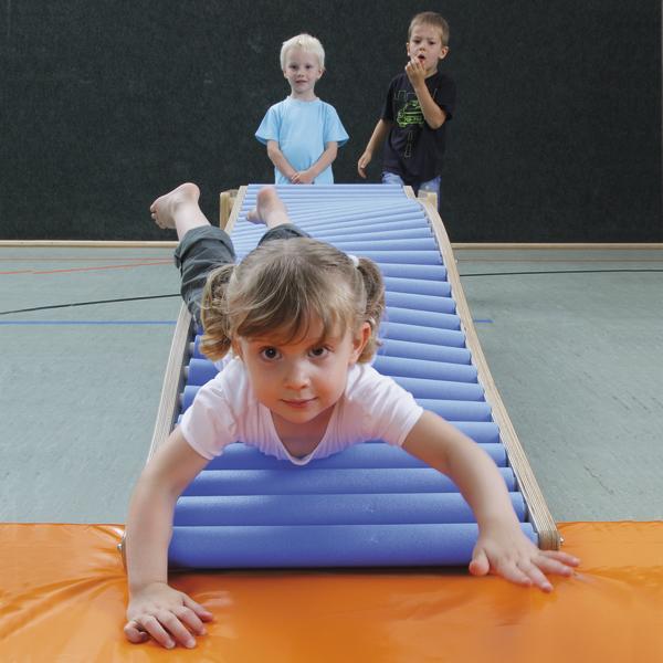 Kind rutscht auf blauer Rolle im Turnraum, zwei Kinder warten im Hintergrund.