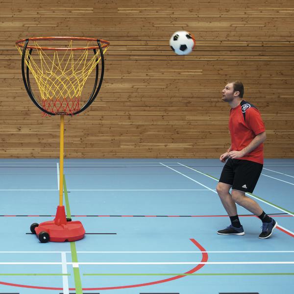 Mann spielt Fußball mit Korbballkorb in Sporthalle, Ball fliegt zum Korb.