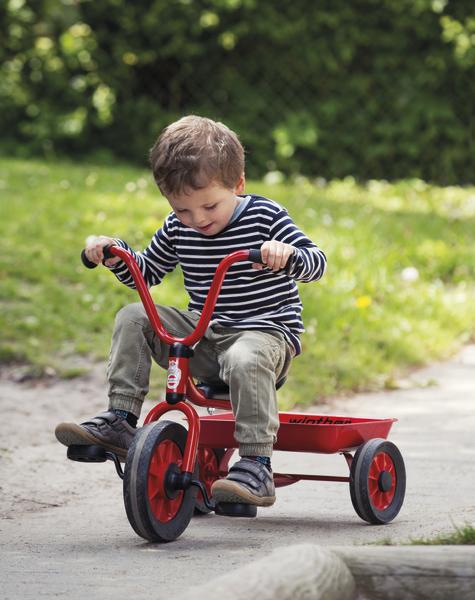 Kind auf rotem Dreirad fährt draußen auf einem Weg im Park bei Sonnenschein.