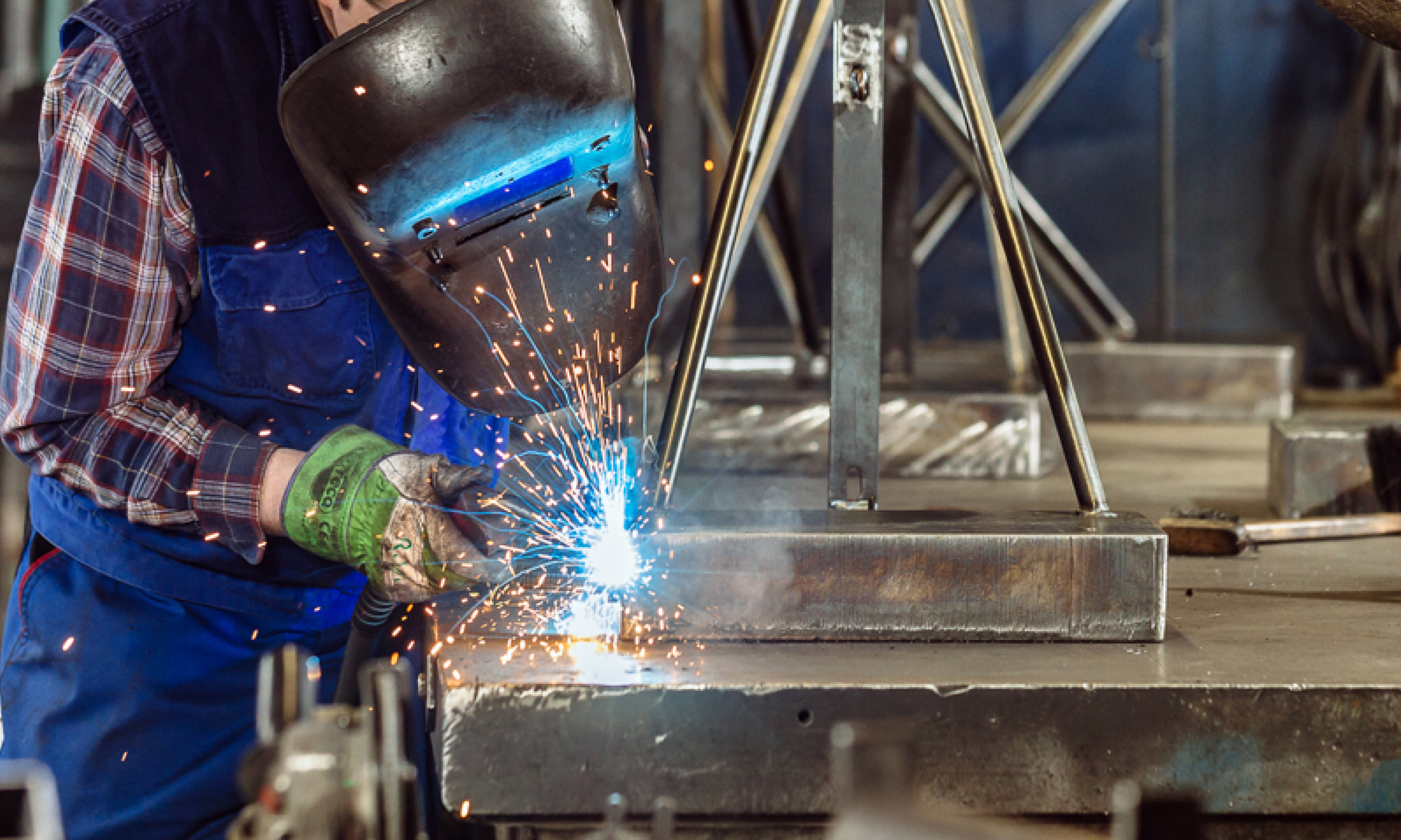 Mitarbeiter mit Schutzhelm schweißt Metallteile auf einem Metalltisch, Funken fliegen beim Schweißvorgang.