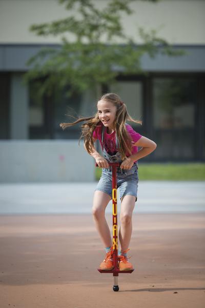 Mädchen hüpft fröhlich mit rotem Pogo-Stick auf glattem Boden im Freien.