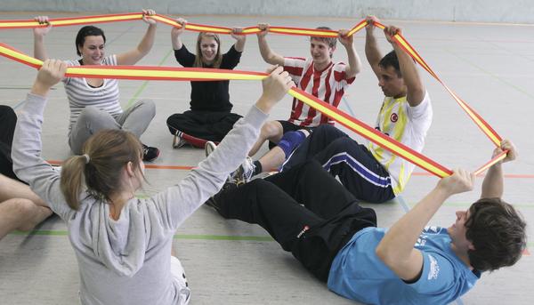 Gruppe von jungen Erwachsenen macht gemeinsam Dehnübungen mit Widerstandsbändern in der Sporthalle.