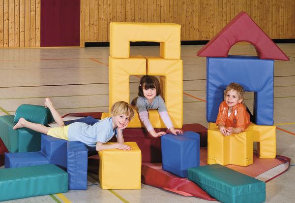 Drei Kinder spielen mit bunten Schaumstoffbausteinen in einer Turnhalle.