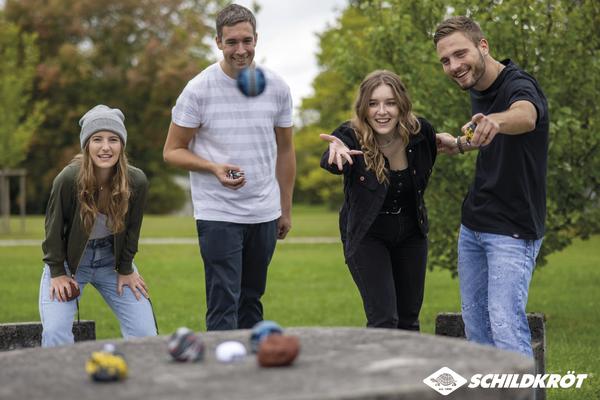 Vier junge Erwachsene spielen draußen Boccia auf einer Betonplatte im Park.