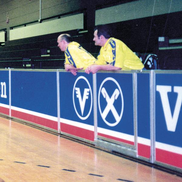 Zwei Männer in gelben Trikots lehnen an der Bande einer Sporthalle während eines Spiels.