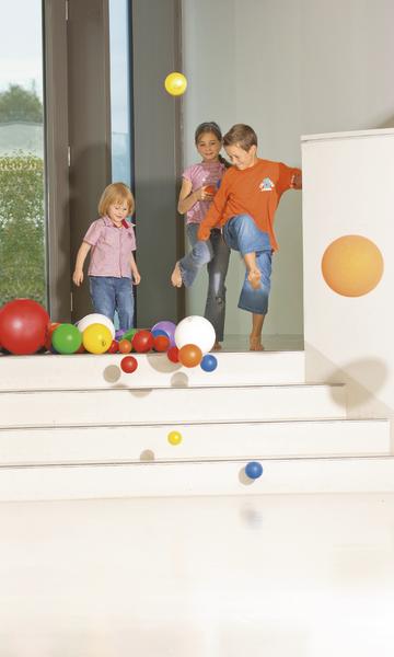 Drei Kinder spielen mit bunten Bällen auf weißen Treppen in hellem Raum.