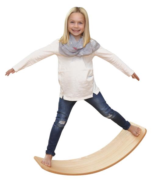 Mädchen balanciert lächelnd auf einem Holz-Wippbrett, trägt Jeans und weißen Pullover.