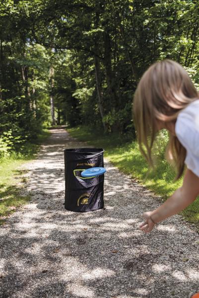 Frau wirft Frisbee in einen Korb auf einem Waldweg bei sonnigem Wetter.