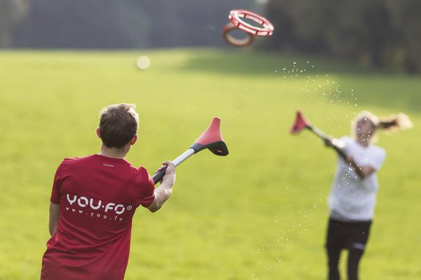Junge wirft Frisbee mit Wurfgerät auf grünem Feld, andere Person fängt es.