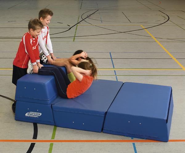 Kinder machen Sit-ups auf blauen Turnmatten in einer Sporthalle mit zwei Helfern.