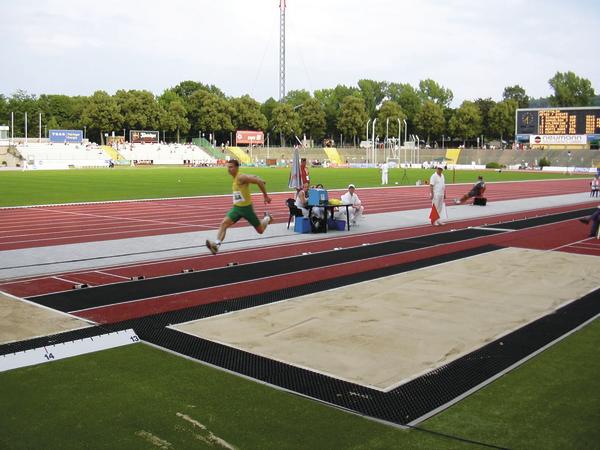 Leichtathlet springt beim Weitsprungwettbewerb in einem Stadion mit Laufbahn und Zuschauern.